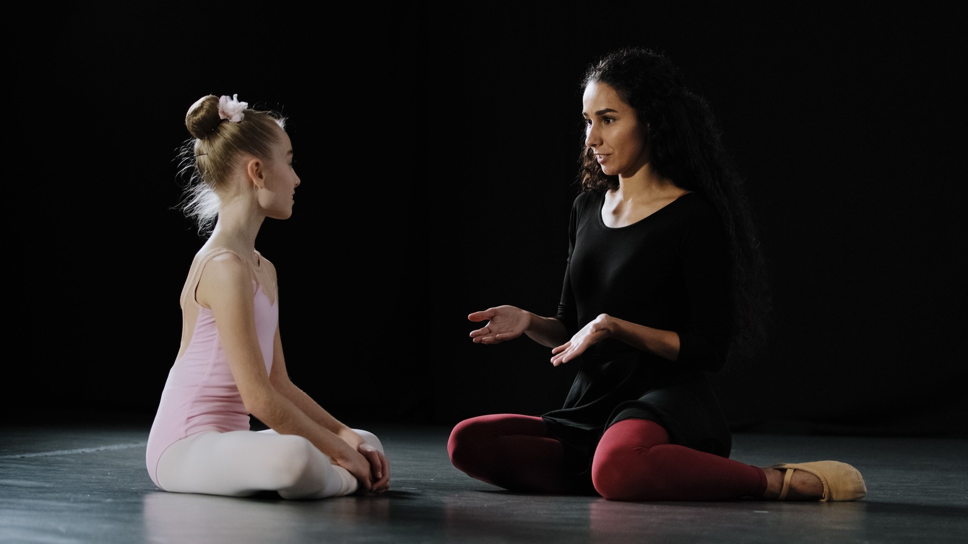 Little girl ballerina dancer beginner acrobat child in leotard sits on floor in dance class with adult woman teacher trainer listening advice talks help training explanation dancing ballet education